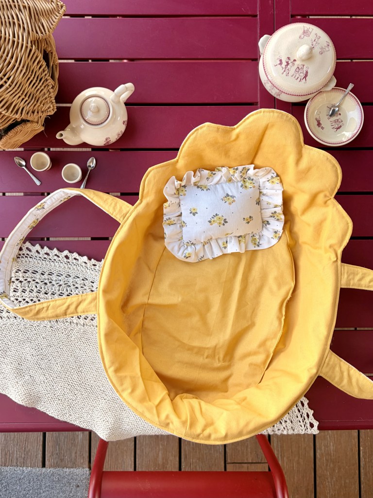 Un panier jaune avec un coussin à volants, placé sur une table en bois, entouré d'une théière, tasses et cuillères.