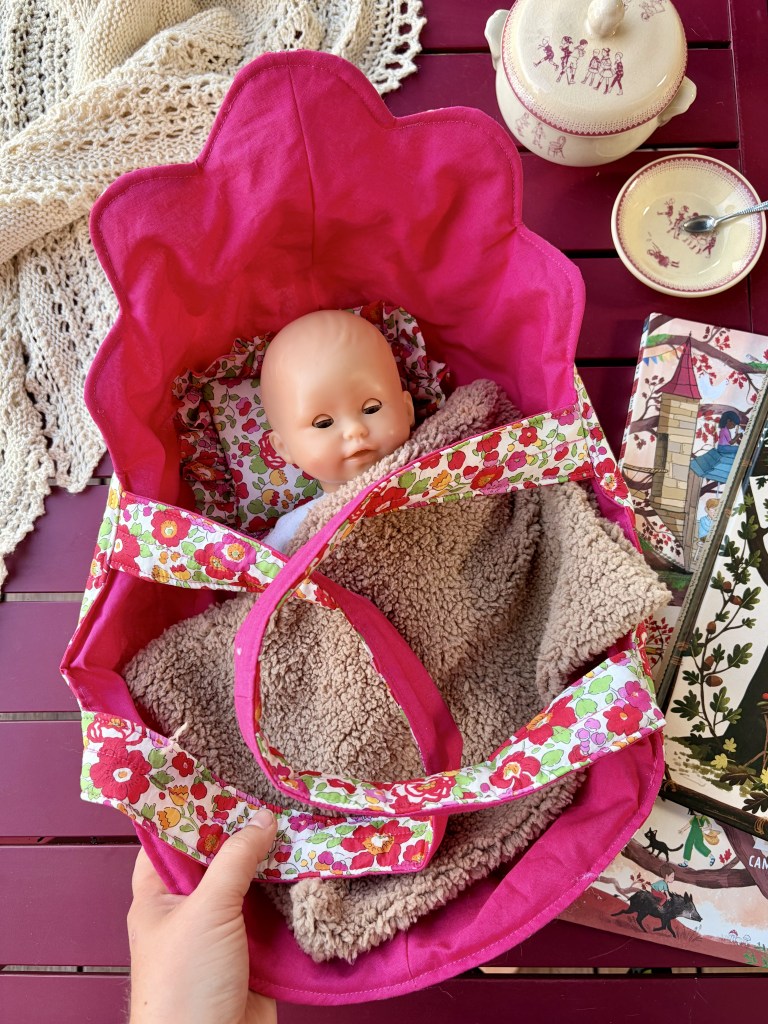 Un panier rose vif contenant une poupée en plastique avec un visage souriant et un intérieur en tissu douillet, posé sur une table en bois.
