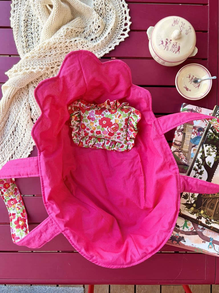 Un sac en tissu rose vif avec des motifs floraux à l'intérieur, posé sur une table en bois, entouré d'une nappe en crochet, d'une théière et de tasses.