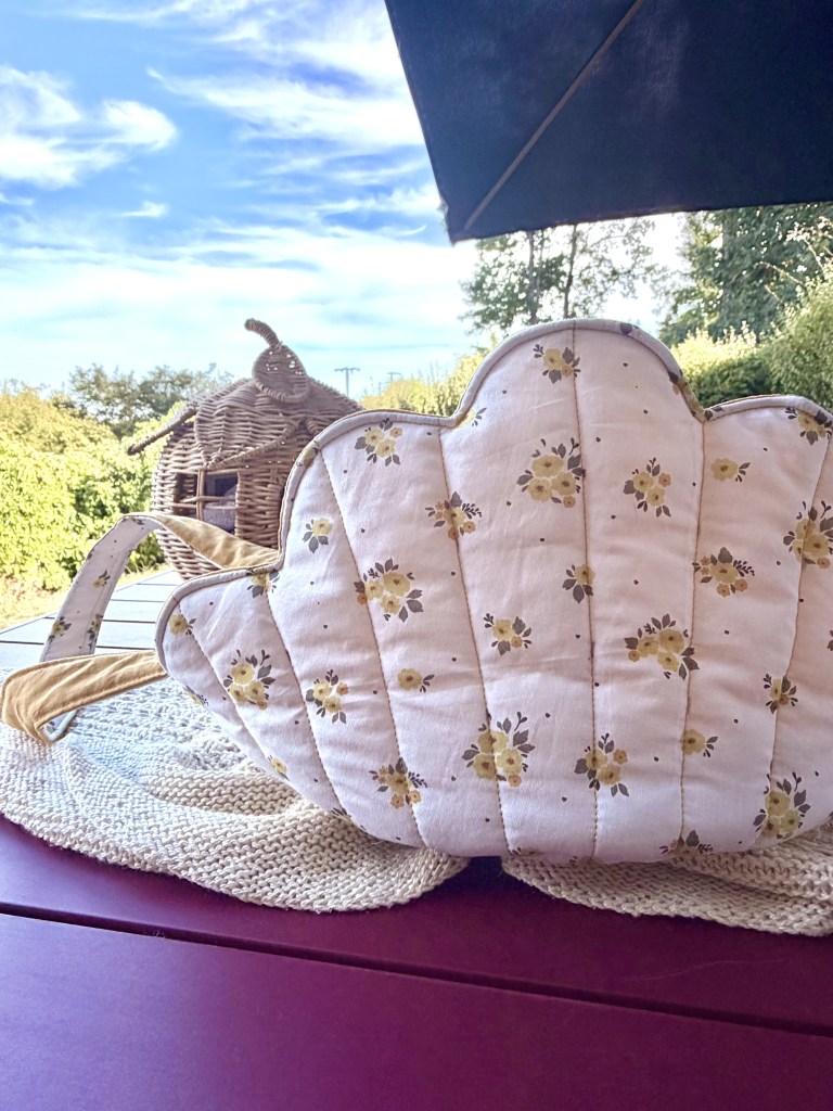 Un sac à main en tissu avec un motif floral, posé sur une nappe en tricot, avec un ciel bleu et des arbres en arrière-plan.