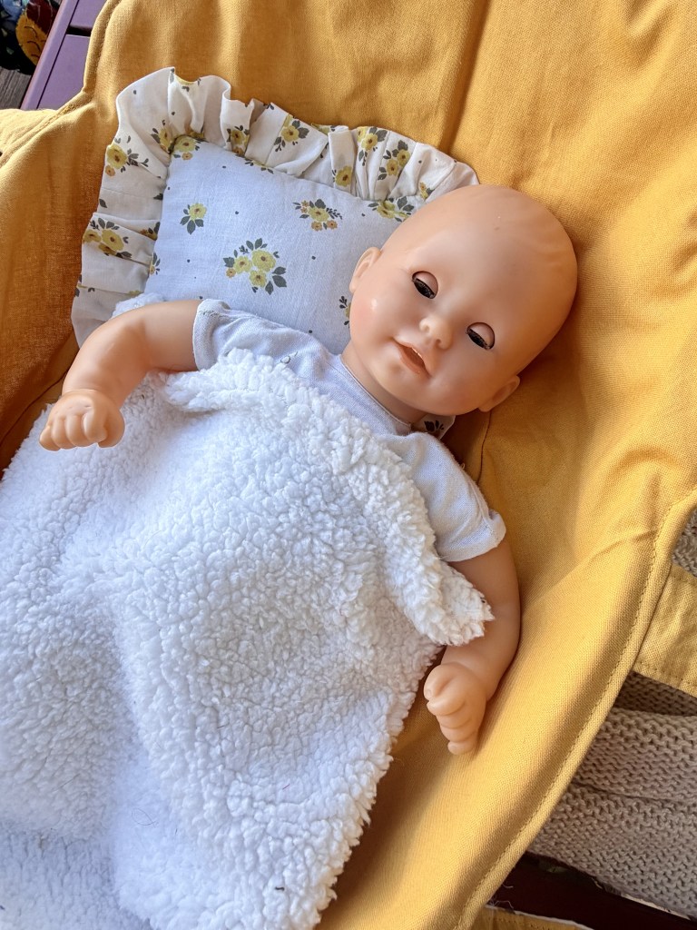 Une poupée allongée dans un berceau, recouverte d'une couverture blanche en peluche, avec un oreiller à motifs floraux.