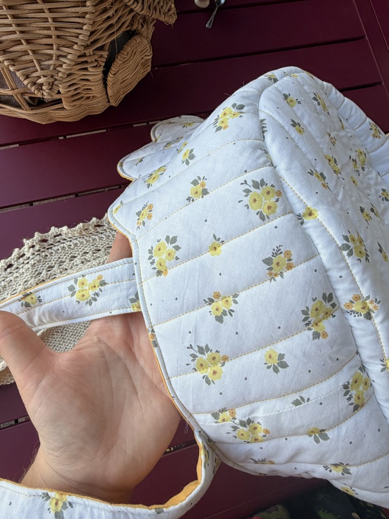 Une main tenant un sac de style patchwork orné de fleurs jaunes sur un fond clair, avec des anses, sur une table en bois.