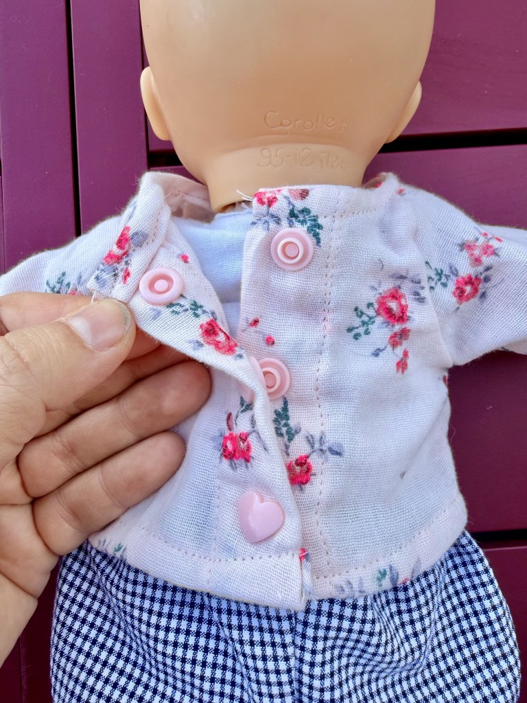 Une main tient une poupée avec une veste à fleurs et des boutons roses, vue de dos.