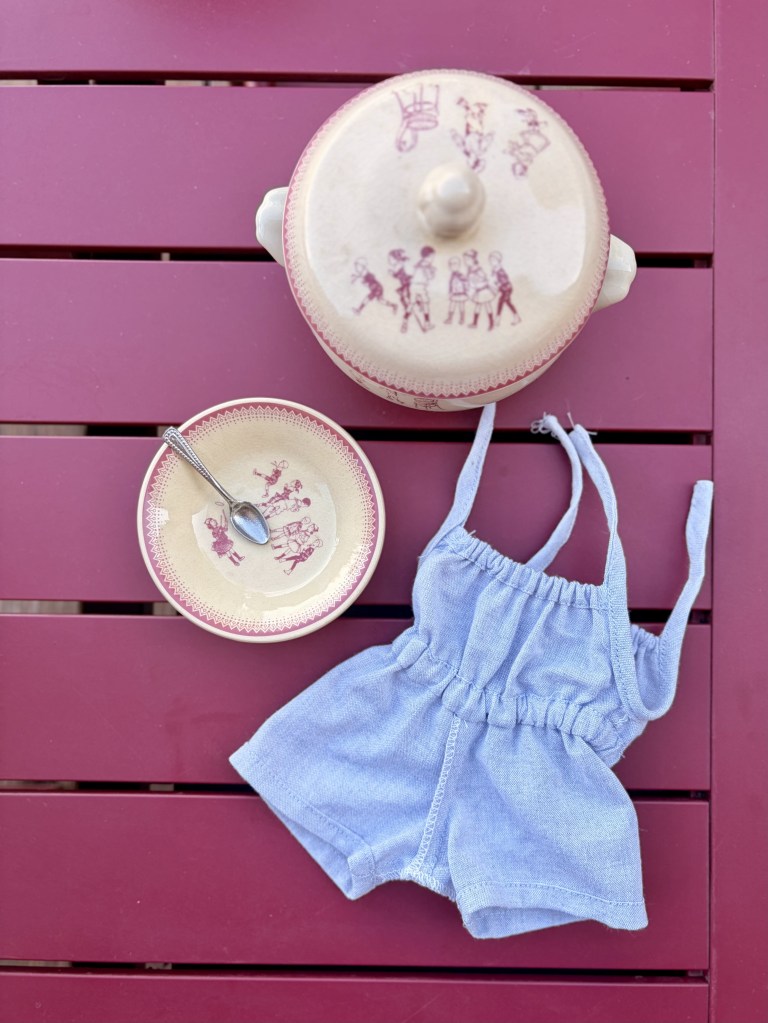 Un ensemble de vêtements en denim bleu, un plat en céramique avec un dessin d'enfants et une cuillère, posés sur une table en bois rouge.