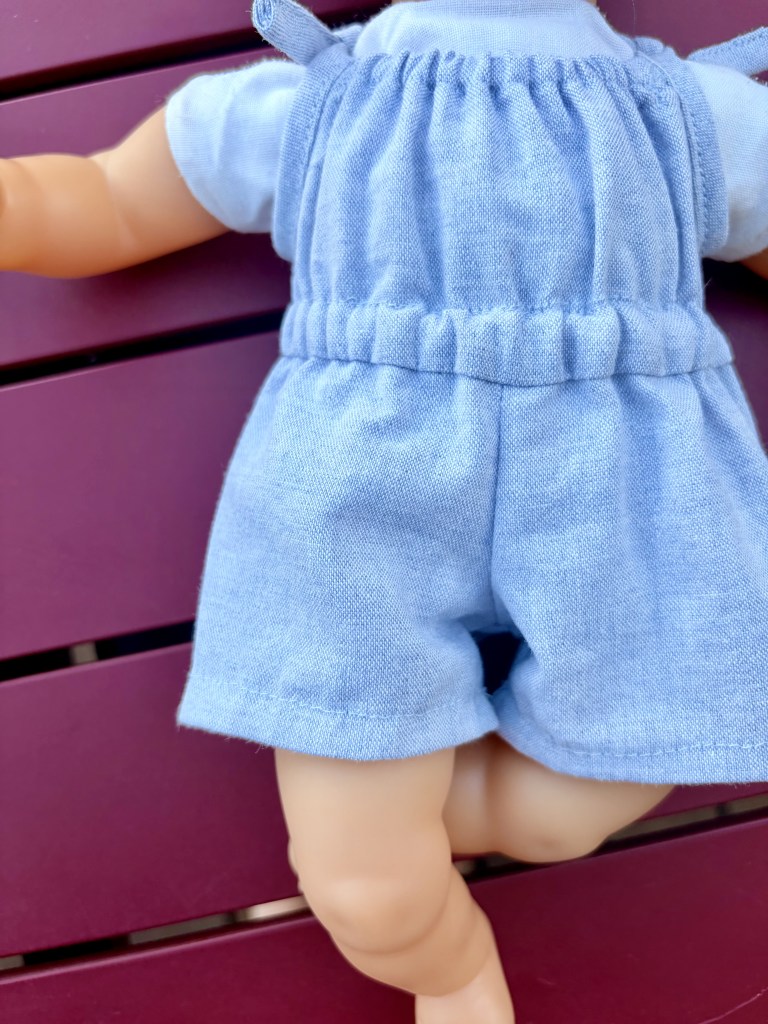 Un gros plan du dos d'une poupée habillée d'une salopette en jean bleu, avec un fond en bois de couleur violette.