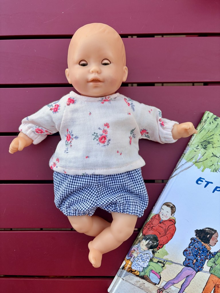 Une poupée en plastique allongée sur une table, portant un haut à fleurs et un short à carreaux, à côté d'un livre illustré.