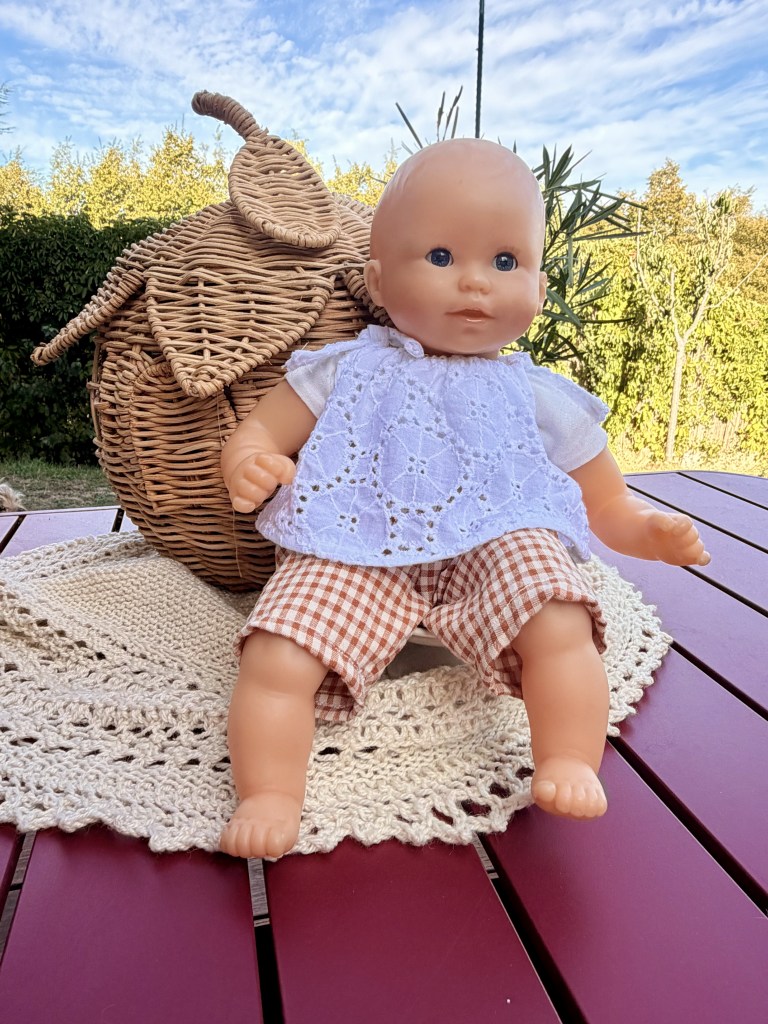 Une poupée assise sur une table avec un panier tressé en arrière-plan, entourée de verdure et d'un napperon.