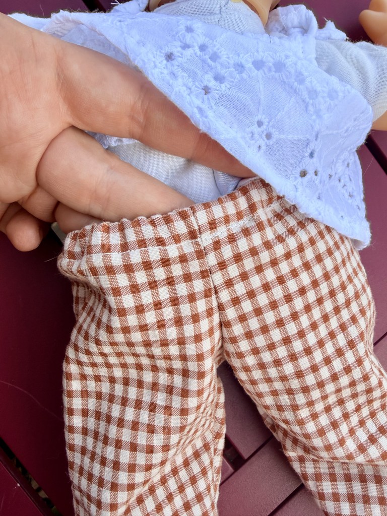 Une main tenant une poupée vêtue d'une blouse bleue et d'un pantalon à carreaux marron.