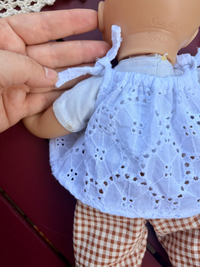 Une main tenant le dos d'une poupée en plastique portant une blouse blanche et un pantalon à carreaux marron.