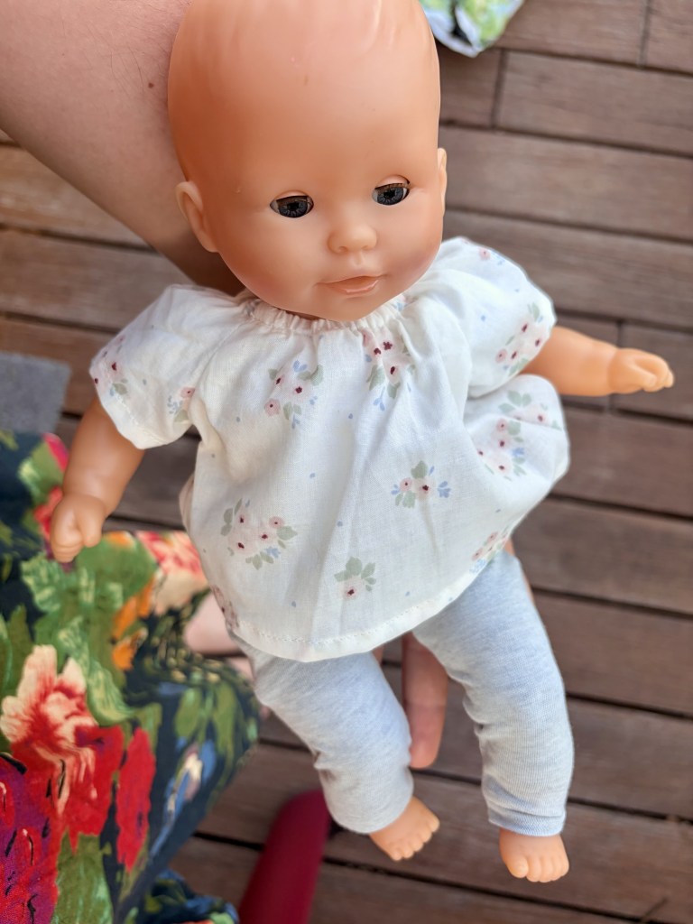 Une poupée en plastique portant un haut à fleurs et un pantalon gris, tenue dans une main.