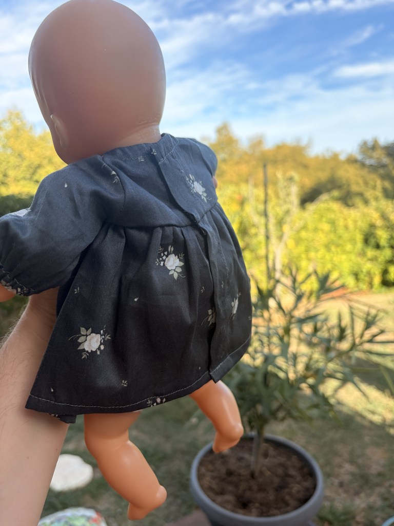 Une poupée en plastique portant une robe à fleurs, tenue par une main, avec un jardin en arrière-plan et un ciel bleu.