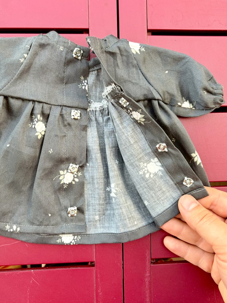 Une petite blouse pour poupée en tissu gris avec des motifs floraux blancs, mise en valeur sur un fond rouge.