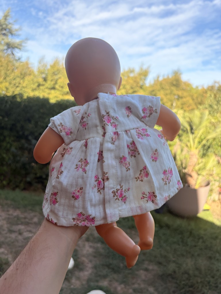 Une main tenant une poupée avec une robe à fleurs, prise en extérieur avec un ciel bleu en arrière-plan.