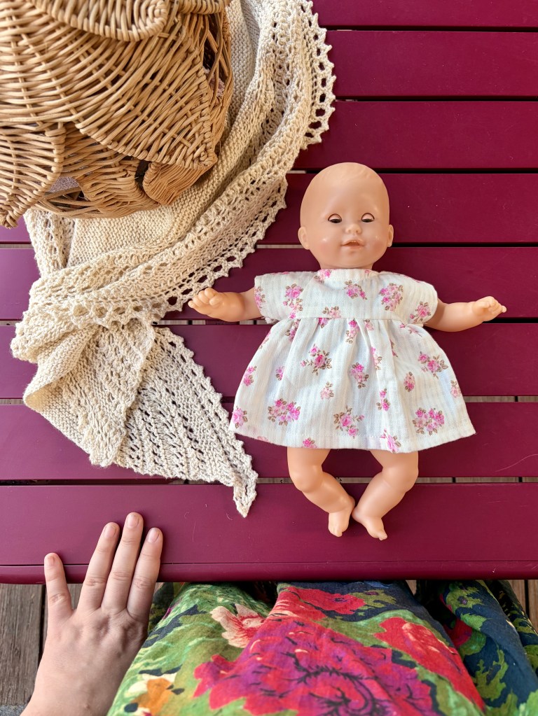 Une poupée en plastique avec une robe à fleurs, posée sur une table rouge, à côté d'un chapeau en osier et d'une écharpe en tricot. La main d'une personne est visible au bas de l'image, avec un tissu coloré en arrière-plan.
