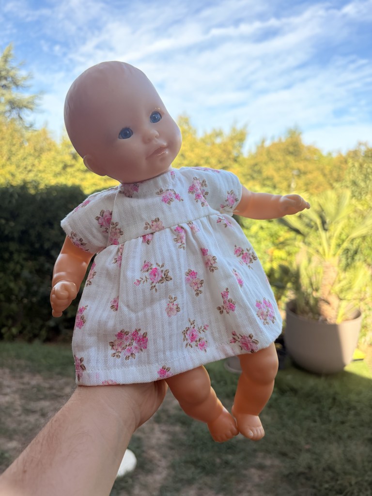 Une poupée en plastique tenant dans une main, portant une robe à fleurs, avec un ciel bleu en arrière-plan.