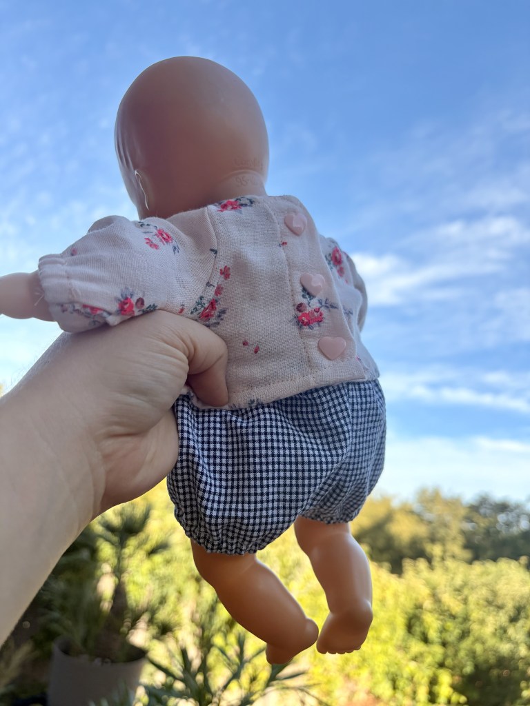 Une main tenant une poupée en plastique, vue de dos, avec un ciel bleu en arrière-plan. La poupée porte une blouse à fleurs et un short à carreaux.